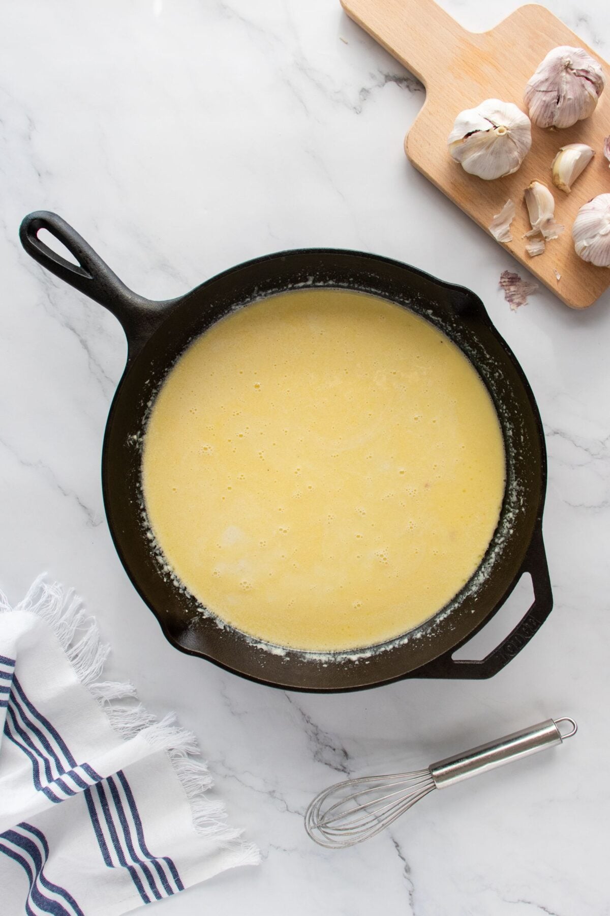 A cast iron skillet filled with a creamy yellow sauce sits on a marble countertop. Nearby are a wooden cutting board with garlic bulbs, a whisk, and a white kitchen towel with dark blue stripes.