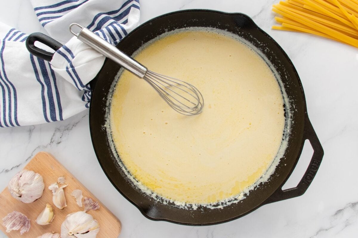 A black skillet with creamy sauce and a whisk inside sits on a marble counter. Uncooked pasta, garlic cloves on a wooden board, and a striped kitchen towel are nearby.