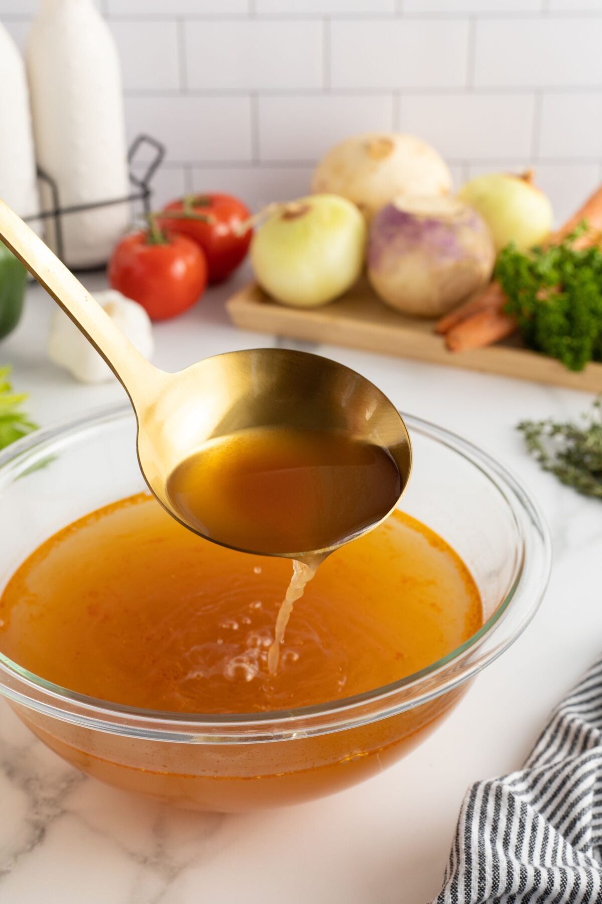 A gold ladle pours broth into a clear glass bowl on a kitchen counter. In the background, onions, turnips, carrots, tomatoes, and fresh herbs rest on a wooden board.