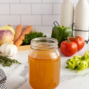 A glass jar filled with vegetable broth sits on a marble counter, surrounded by fresh vegetables like tomatoes, carrots, garlic, bell pepper, celery, and herbs, with kitchen items in the background.