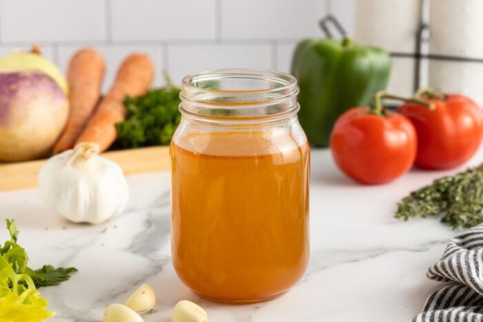 A glass mason jar filled with golden vegetable broth sits on a white countertop, surrounded by fresh vegetables including tomatoes, a green bell pepper, carrots, garlic, and leafy greens.