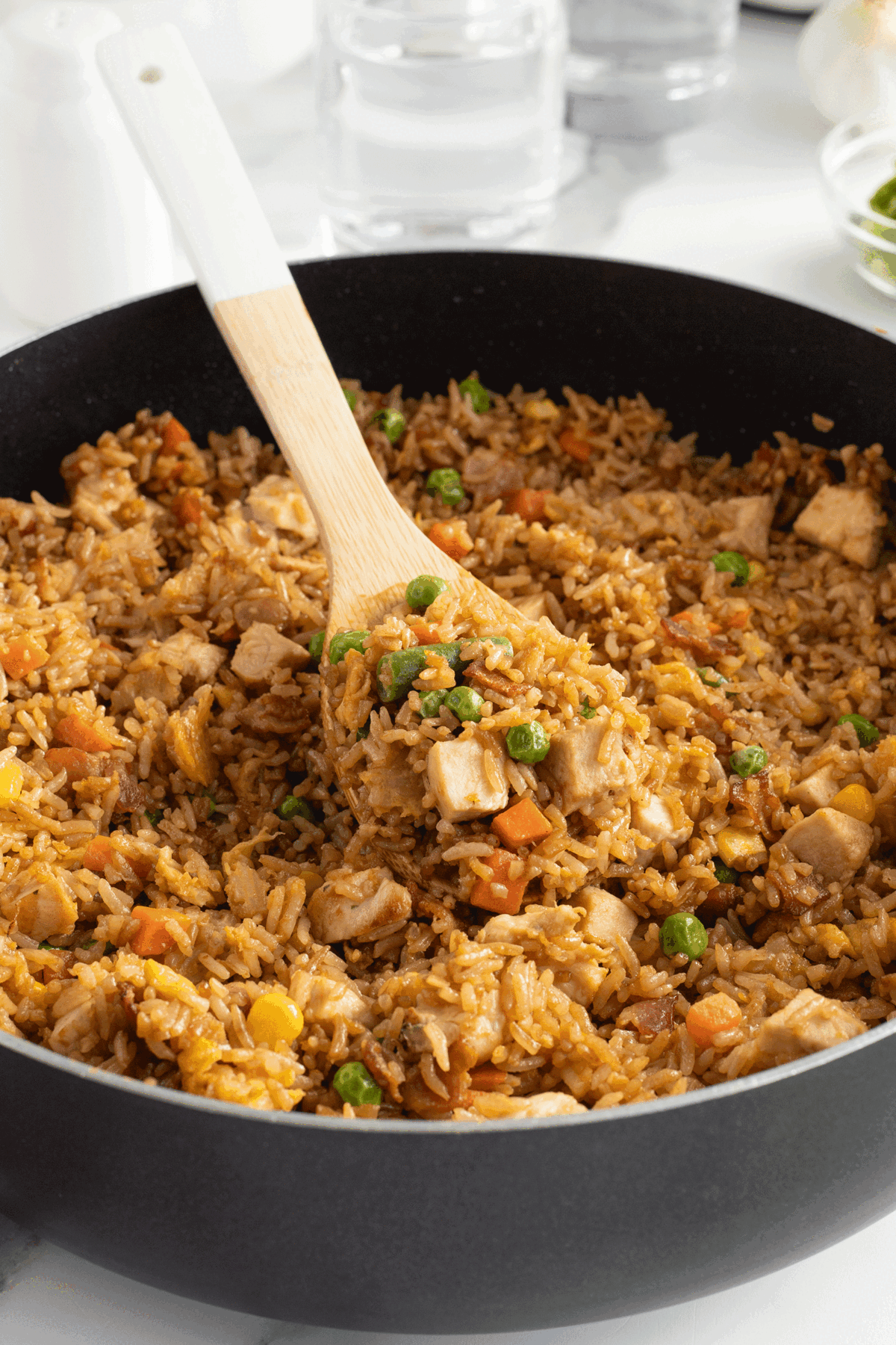 A wooden spoon scoops a portion of fried rice with diced tofu, peas, corn, and carrots from a large black pan. Glasses of water and condiments are visible in the blurred background.