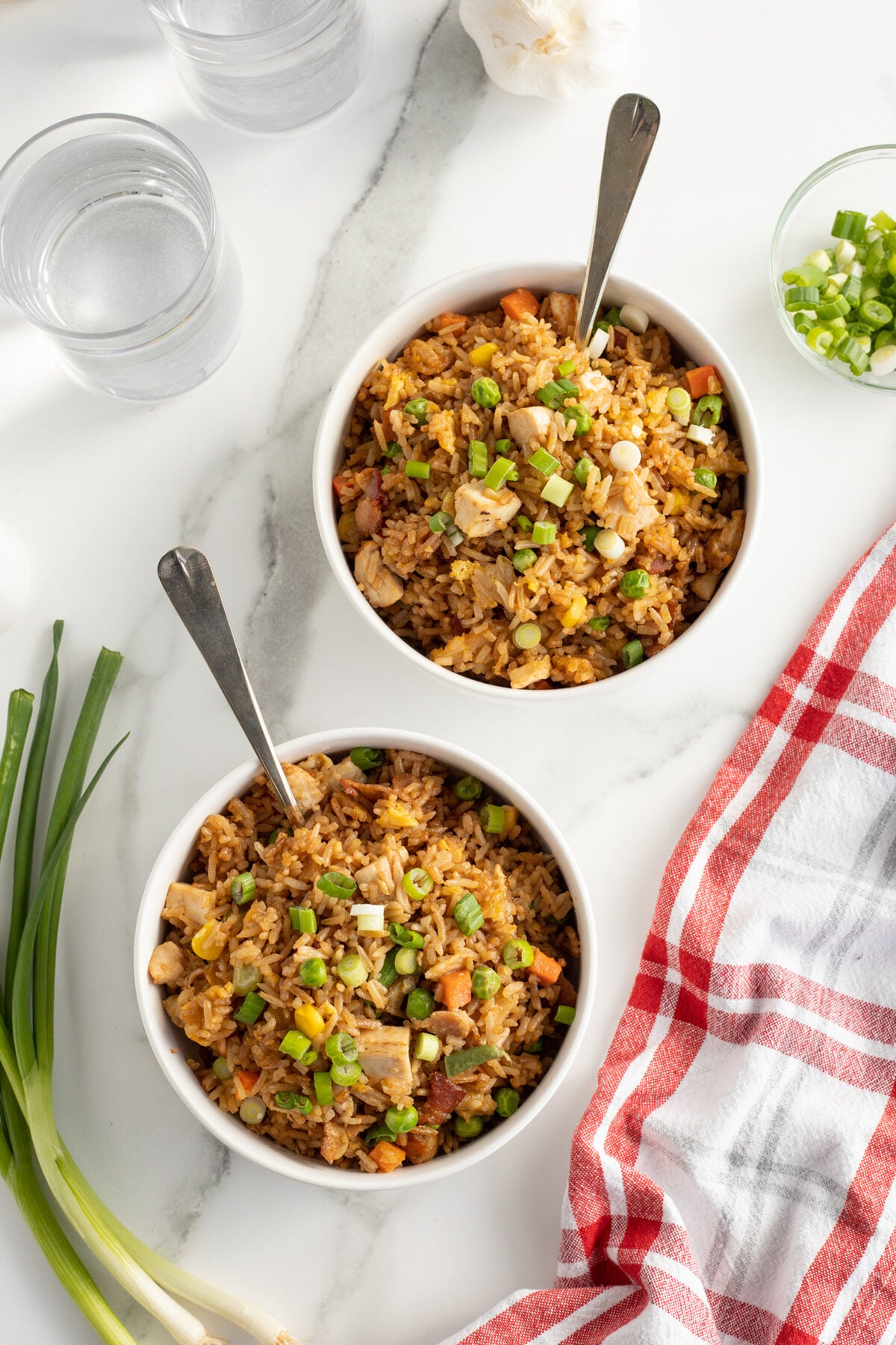 Two bowls of fried rice with vegetables and green onions, each with a spoon, sit on a white marble surface next to glasses of water, fresh green onions, a red-and-white checkered napkin, and a small bowl of chopped green onions.