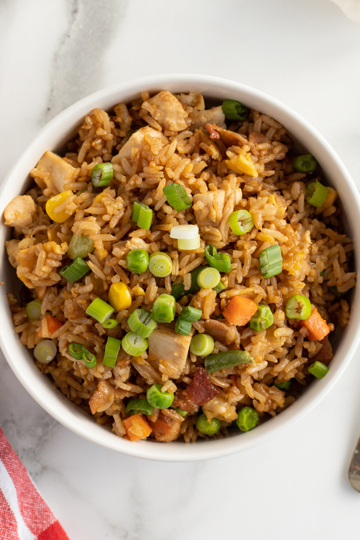 A bowl of fried rice with diced chicken, carrots, corn, green beans, peas, and chopped green onions on top, placed on a white marble surface.