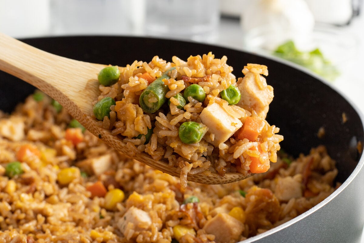 A wooden spoon holds a serving of fried rice with peas, carrots, corn, and tofu over a pan filled with more of the same fried rice.