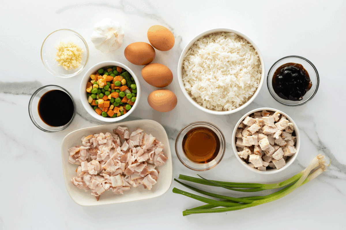 Various ingredients on a white surface, including cooked rice, eggs, diced chicken, chopped bacon, mixed vegetables, sliced green onions, minced garlic, soy sauce, oyster sauce, and a small bowl of oil.
