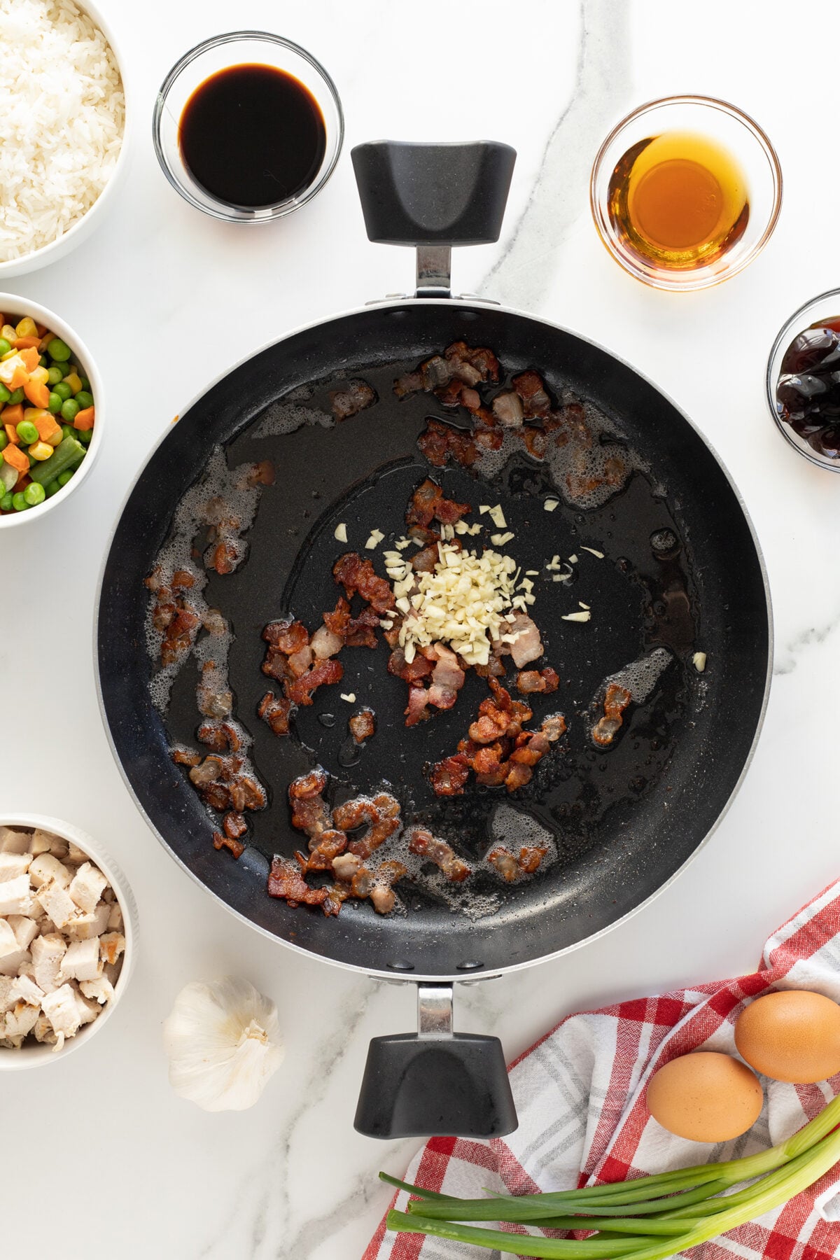 A frying pan with cooked bacon and minced garlic, surrounded by bowls of mixed vegetables, rice, cooked chicken, eggs, sauces, and a garlic bulb on a white countertop with a red and white towel and green onions.