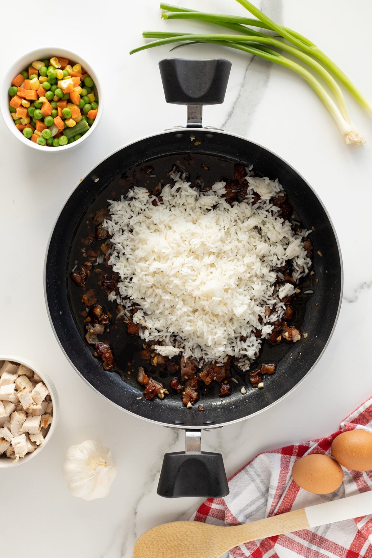 A frying pan with cooked white rice and diced meat sits on a marble counter, surrounded by bowls of mixed vegetables and diced chicken, eggs, garlic, green onions, and a wooden spoon on a red-striped towel.