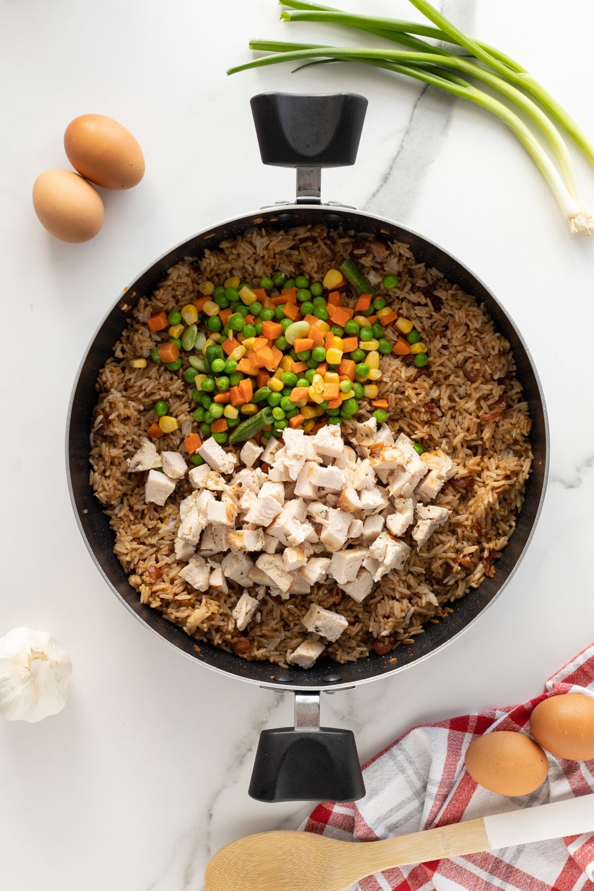 A black skillet contains fried rice topped with diced chicken breast and a mix of peas, carrots, and corn. Surrounding the skillet are two eggs, green onions, garlic, a wooden spoon, and a red-checked towel on a white surface.