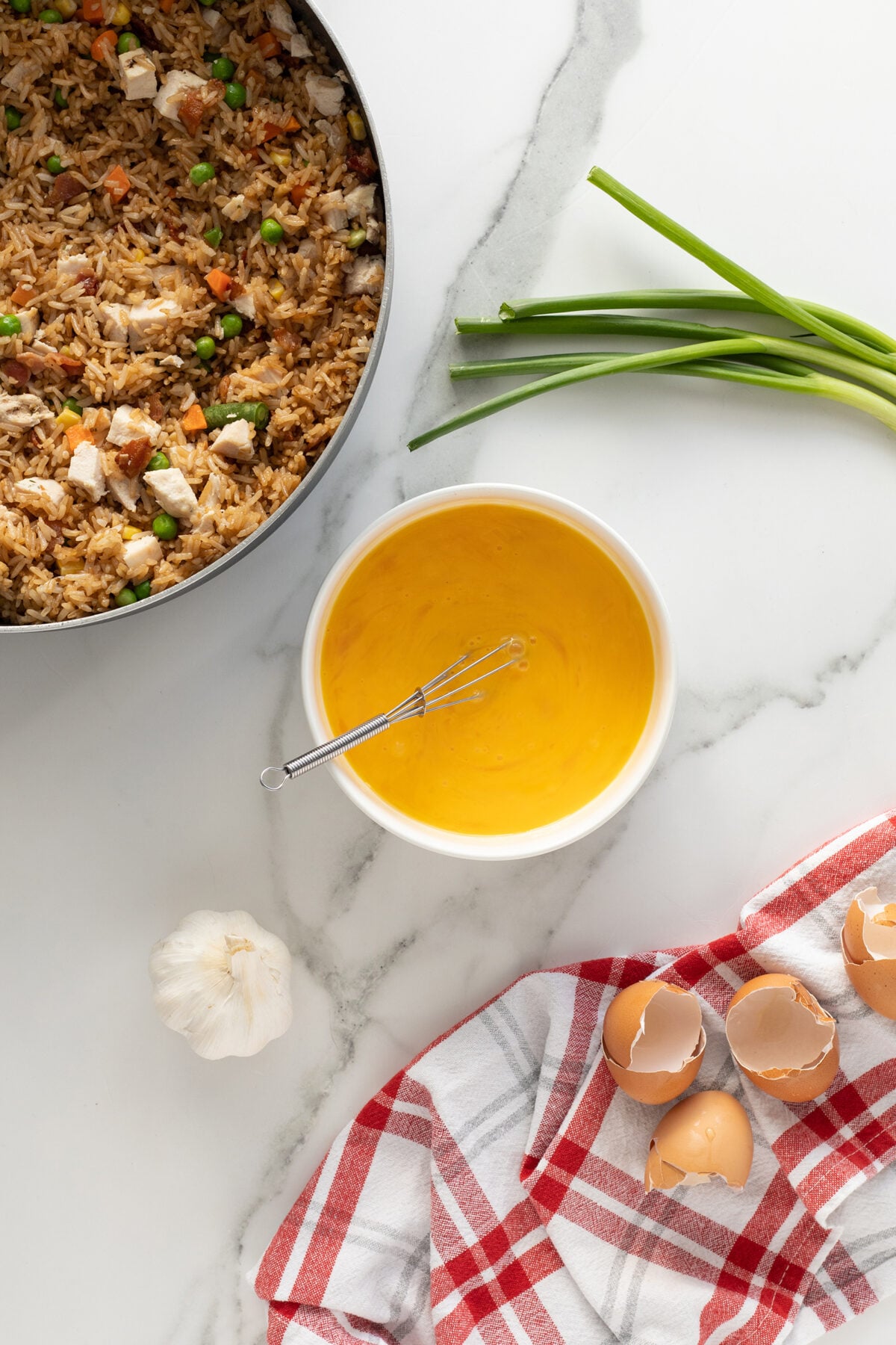 A bowl of beaten eggs with a whisk, cracked eggshells on a red and white towel, a head of garlic, green onions, and a pan of fried rice with vegetables and chicken on a white marble countertop.