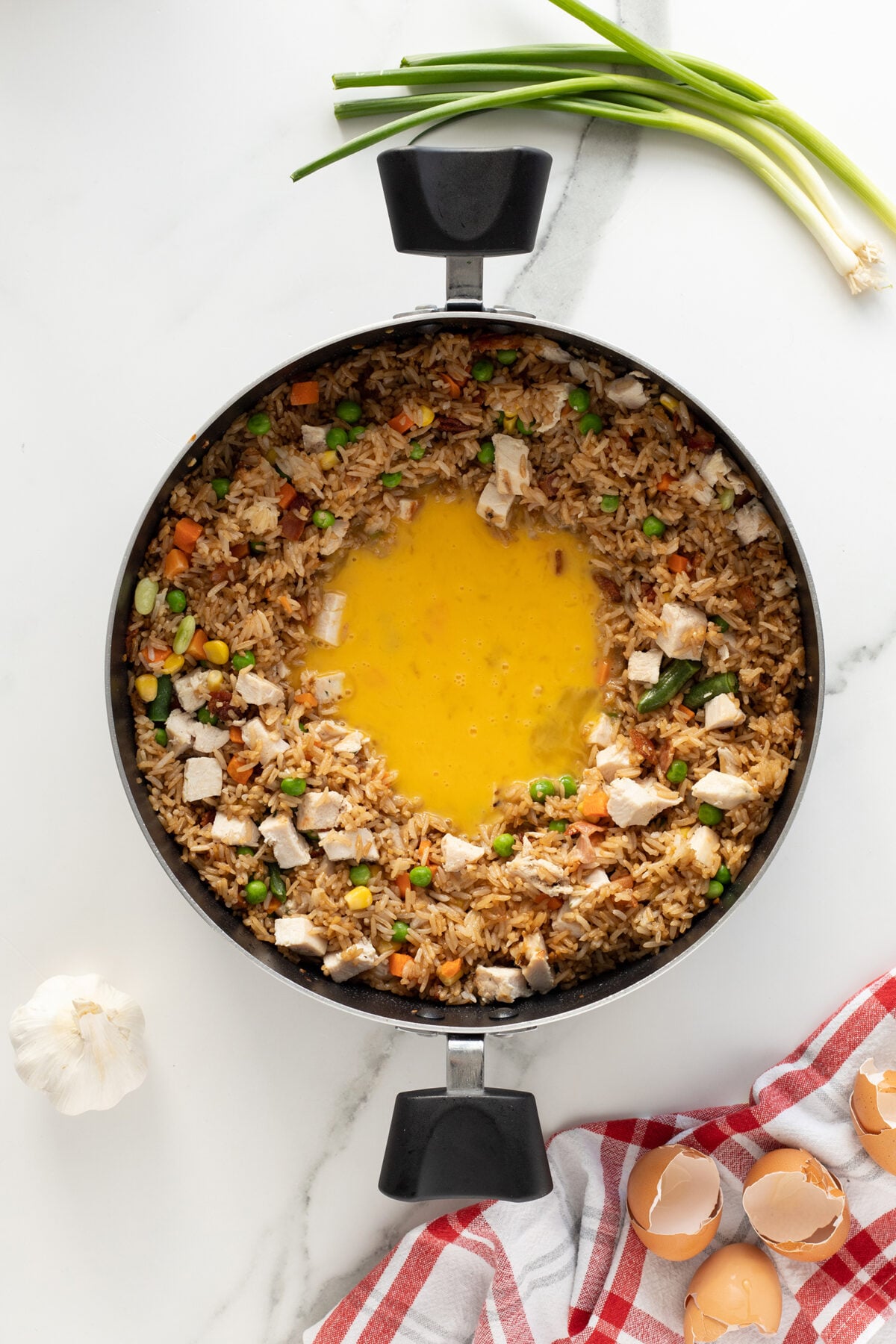 A skillet with fried rice, vegetables, and chicken arranged in a ring, with beaten eggs in the center. Green onions, garlic, and eggshells are on a white marble surface nearby, along with a red-striped towel.