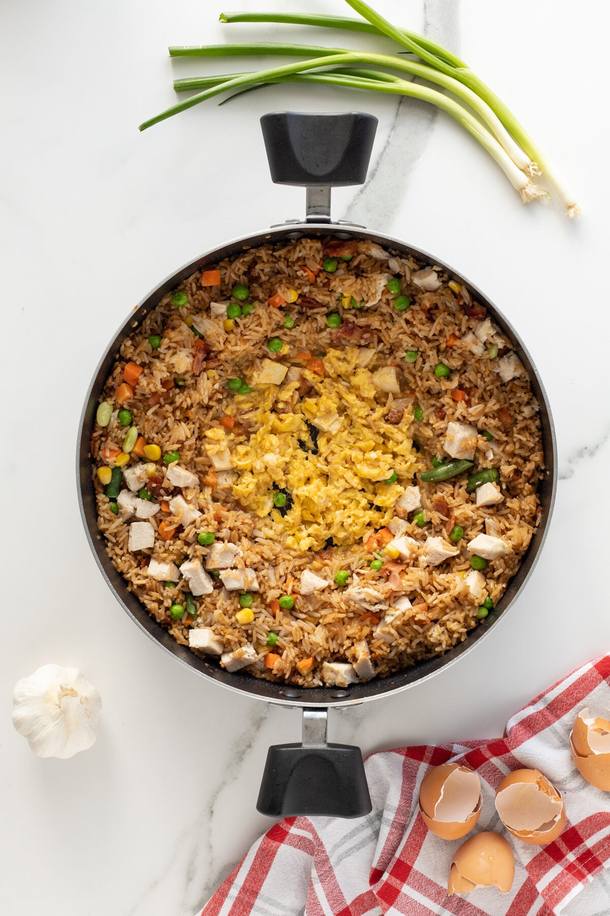 A skillet filled with chicken fried rice topped with scrambled eggs, diced vegetables, and chicken pieces. Nearby are green onions, a bulb of garlic, and cracked eggshells on a red and white cloth.