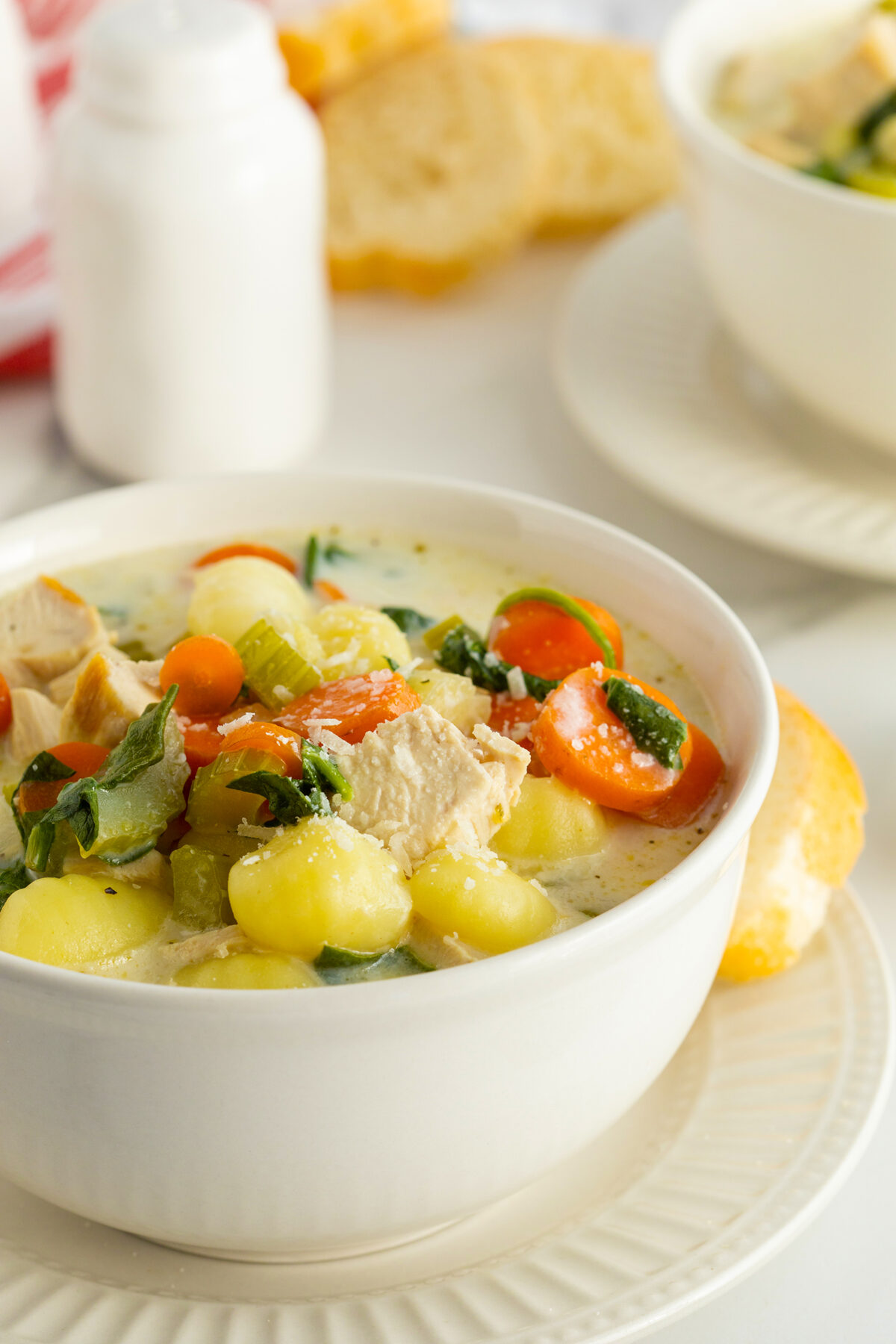 A bowl of creamy chicken and gnocchi soup with pieces of chicken, gnocchi, carrots, celery, and spinach, served with a bread roll on a white plate. Another bowl and bread are in the background.