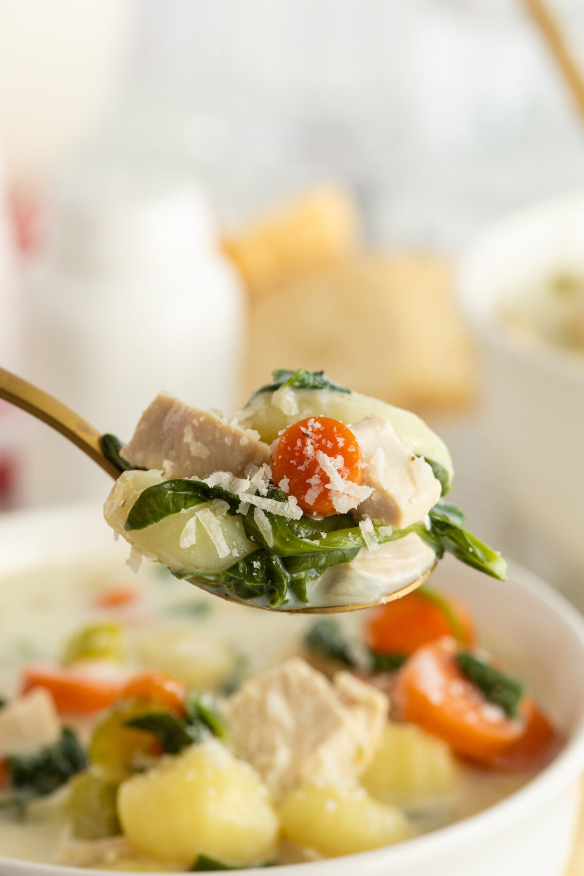 A close-up of a spoonful of creamy chicken and vegetable soup, featuring chunks of chicken, spinach, carrots, potatoes, and grated cheese, held above a bowl of the same soup.