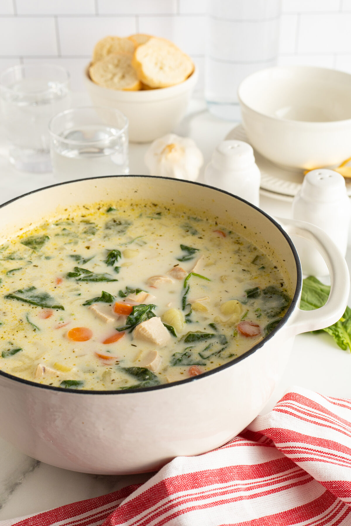A white pot filled with creamy chicken soup containing carrots, spinach, and herbs sits on a countertop near a red-striped towel, with bread, garlic, bowls, glasses of water, and salt and pepper shakers in the background.