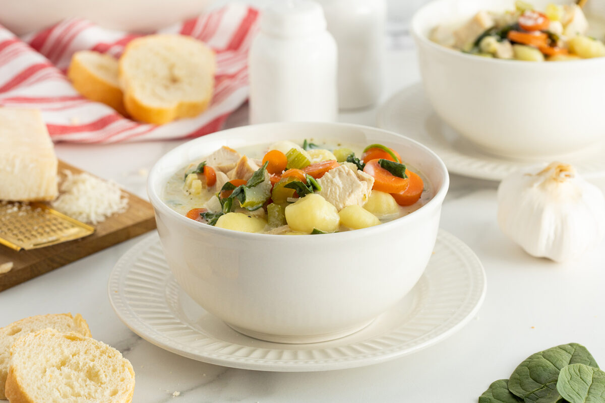 A white bowl of creamy soup with chunks of chicken, gnocchi, carrots, and spinach sits on a white plate. Nearby are slices of bread, a garlic bulb, a cheese grater, and a red-striped towel.