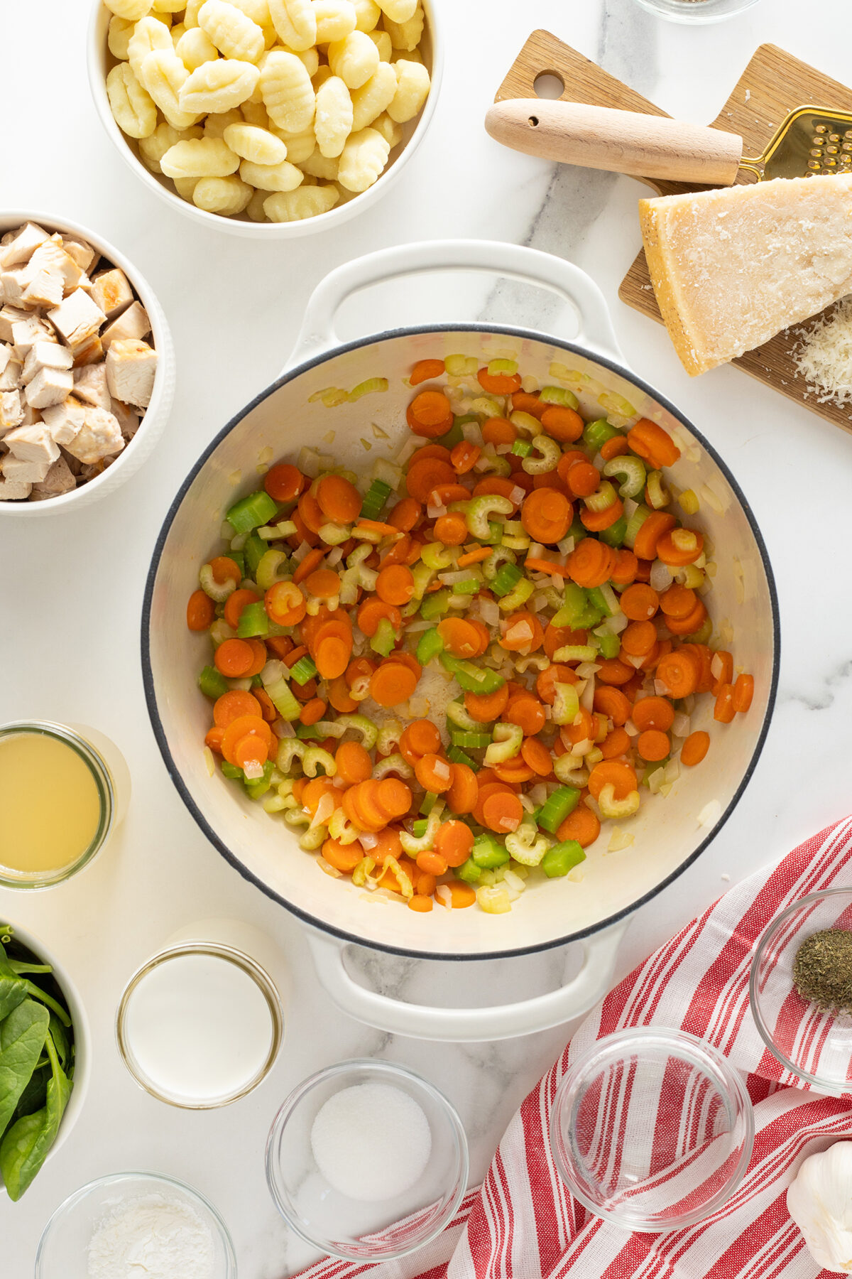 A white pot contains sautéed carrots and celery. Surrounding it are bowls of gnocchi, cubed chicken, spinach, broth, shredded Parmesan, and various spices on a white marble counter with a red-striped dish towel.