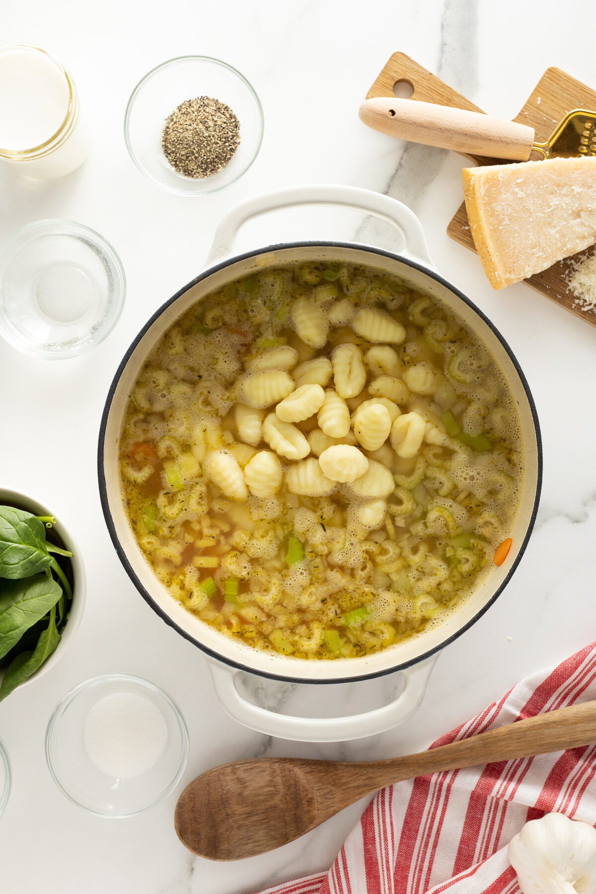 Top view of a pot of soup with gnocchi, vegetables, and broth on a white surface, surrounded by bowls of spinach, seasonings, grated cheese, and a wooden spoon with a red-striped towel.