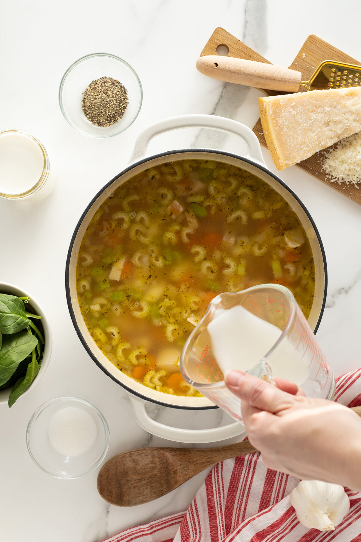 A hand pours milk from a measuring cup into a pot of soup on a white counter, surrounded by a bowl of spinach, grated parmesan, black pepper, garlic, and a red-striped towel.
