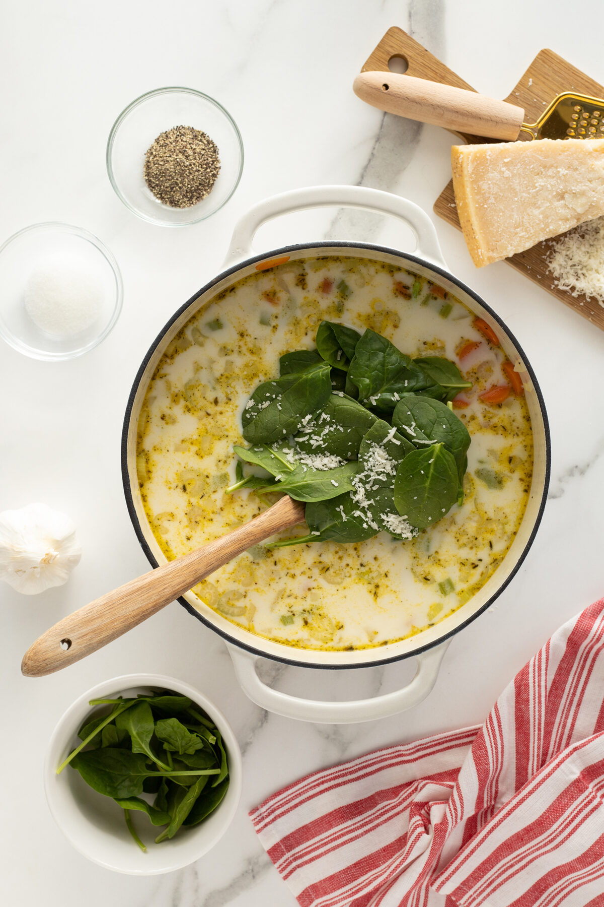 A white pot of creamy soup with fresh spinach and grated cheese on top sits on a marble counter. Nearby are a bowl of spinach, a block of Parmesan cheese, a grater, salt, pepper, garlic, and a red-striped towel.