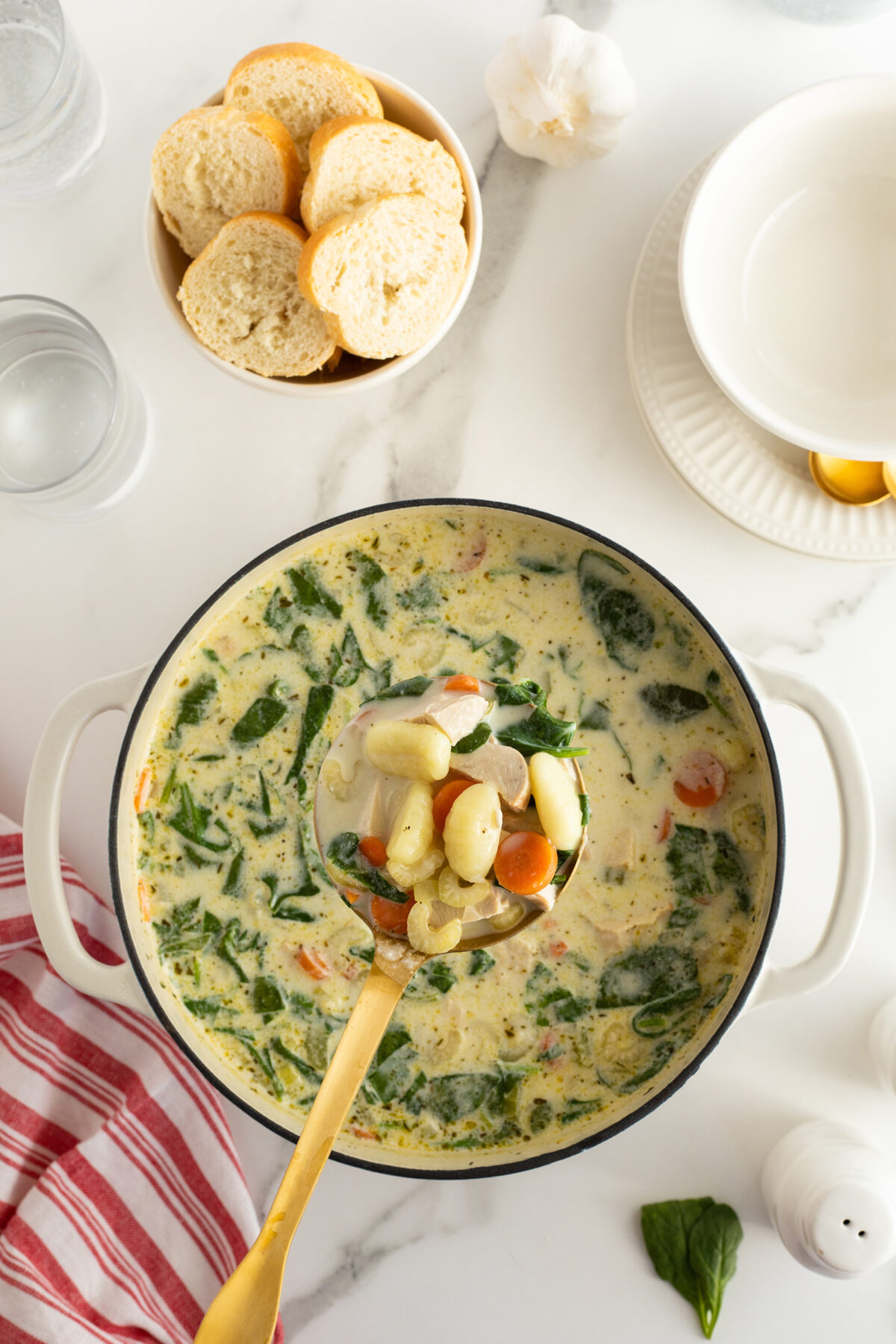 A pot of creamy soup with spinach, gnocchi, and vegetables sits on a marble surface. A ladle holds a serving above the pot. Nearby are sliced bread, a garlic bulb, empty bowls, and a striped red towel.