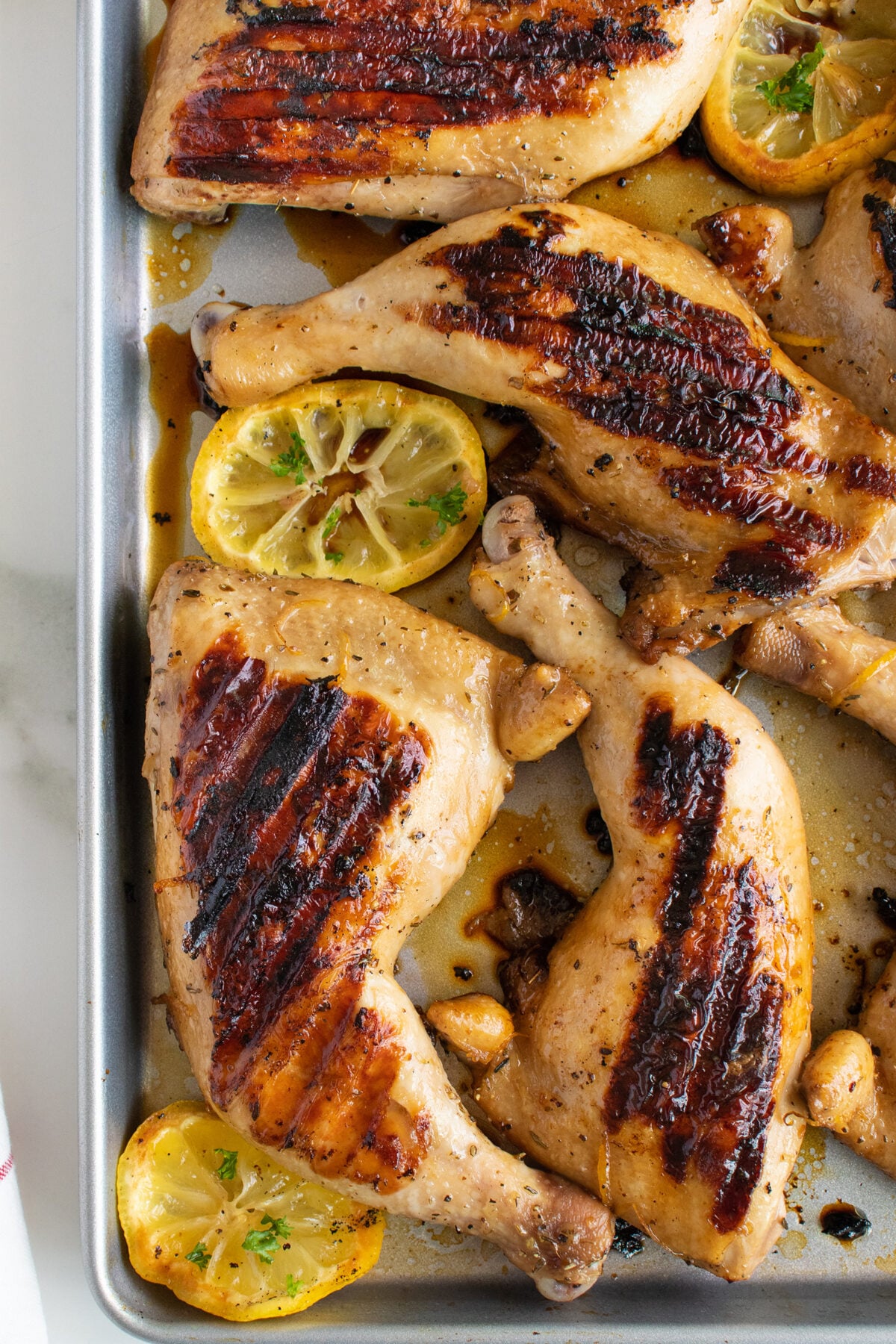 Several grilled chicken leg quarters with char marks are arranged on a baking sheet, garnished with slices of roasted lemon. The chicken appears juicy and well-seasoned.