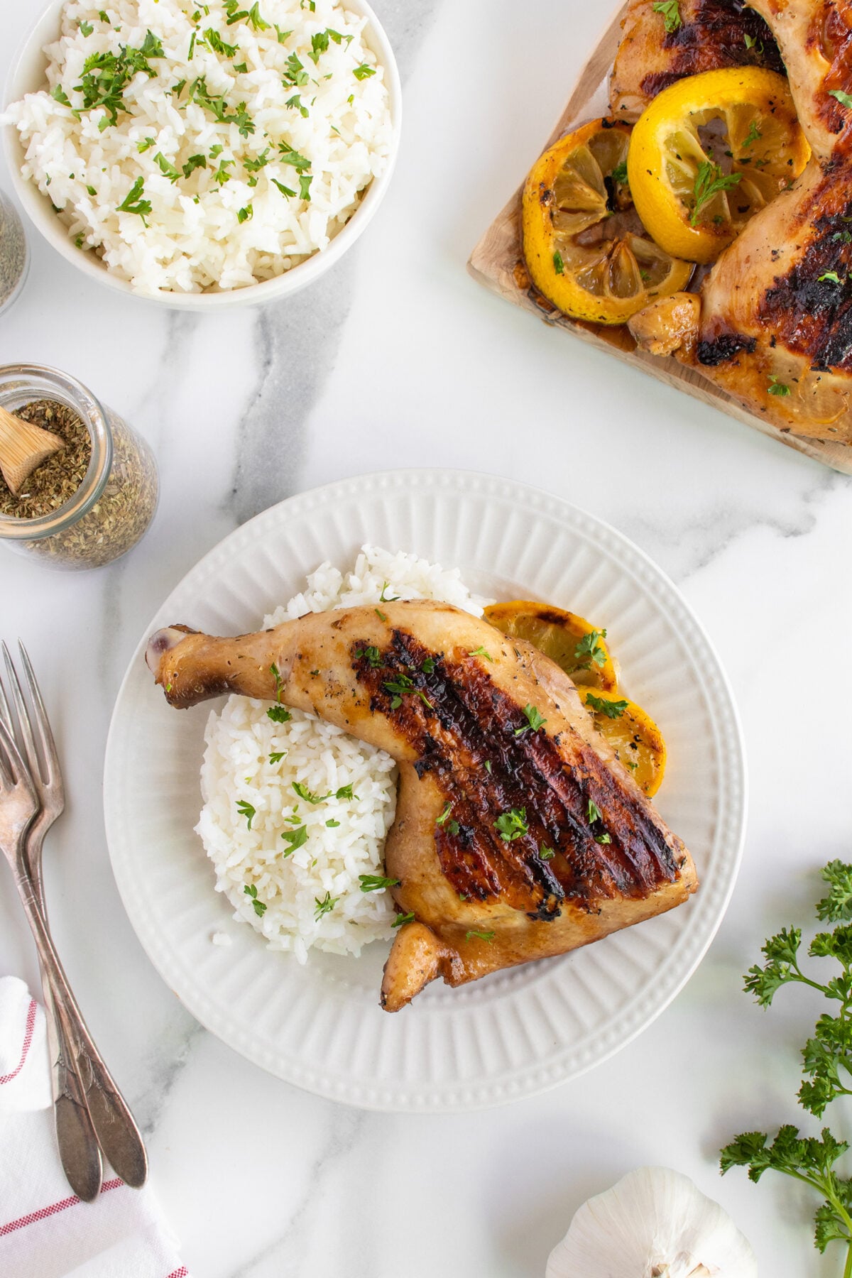 A grilled chicken leg quarter with herbs rests on a bed of white rice on a white plate, surrounded by a bowl of rice, grilled chicken in a dish, seasonings, and fresh parsley on a marble surface.