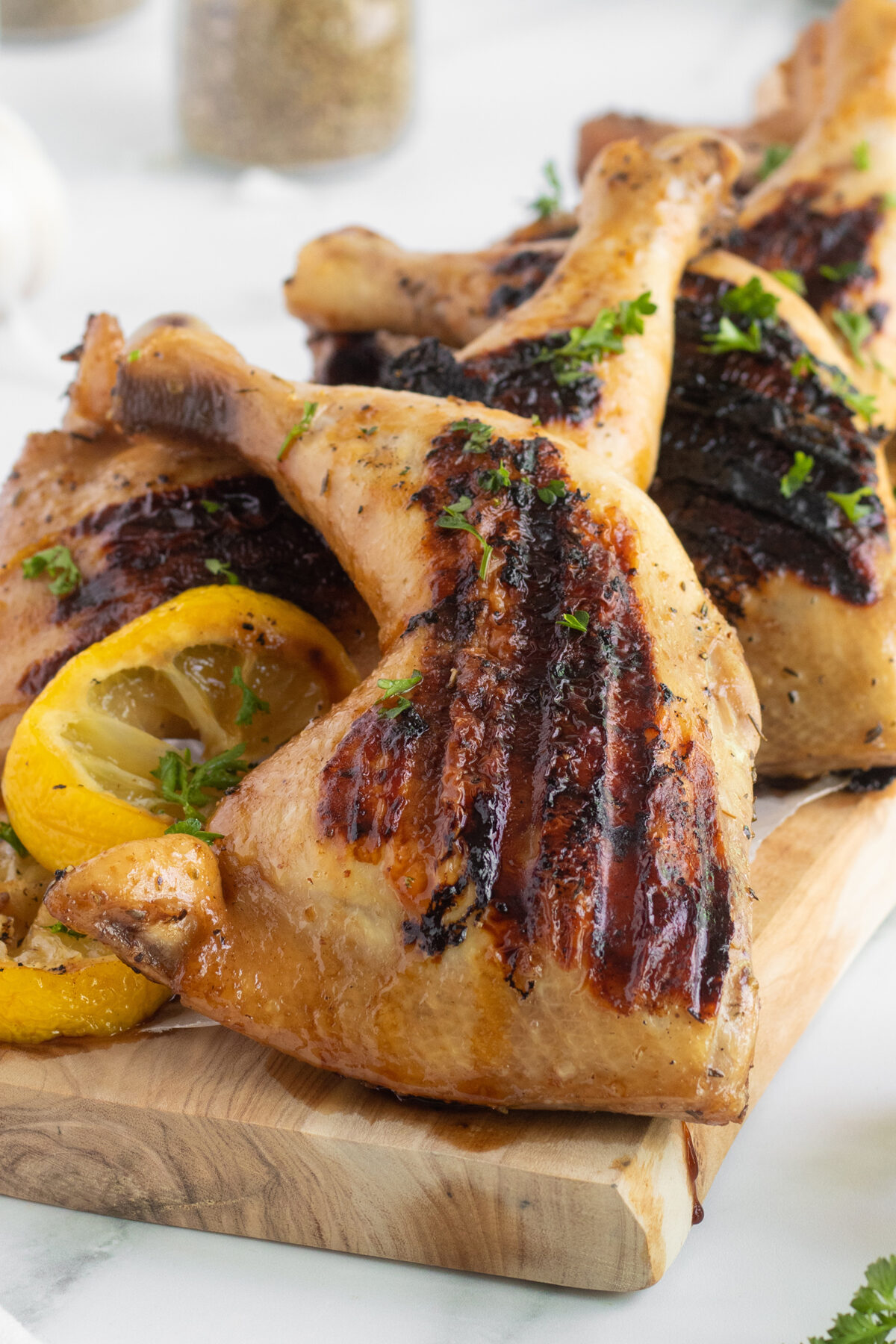 Grilled chicken legs with char marks are arranged on a wooden board, garnished with chopped herbs and lemon slices. The chicken appears juicy and golden-brown, ready to be served.