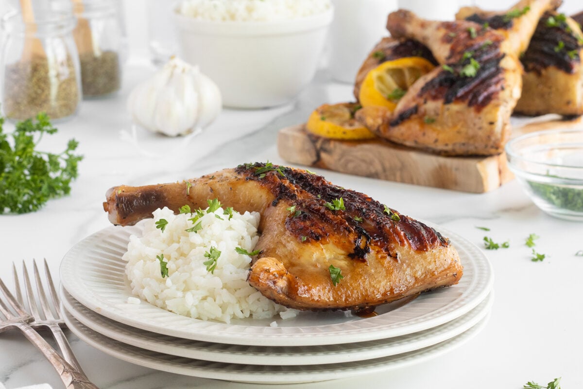 A grilled chicken leg quarter garnished with herbs is served on a plate with white rice. More chicken, rice, garlic, and herbs are visible in the background on a white table.
