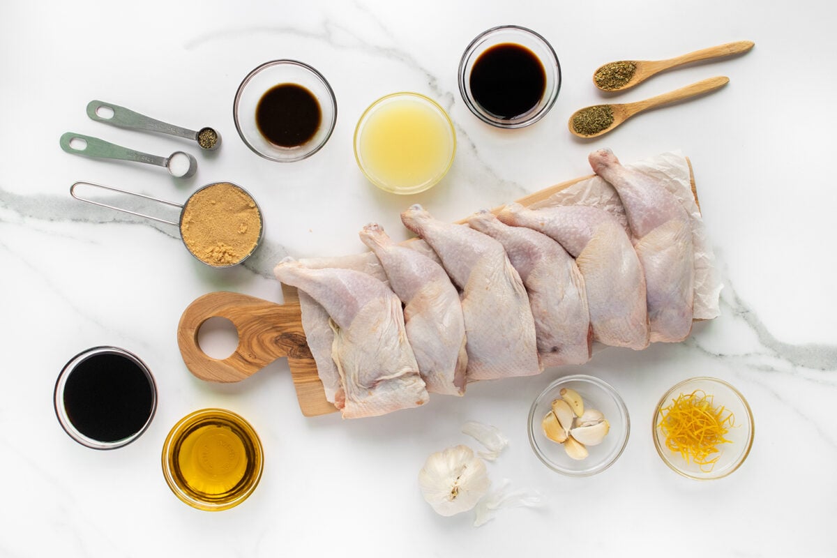 Raw chicken legs arranged on a wooden board surrounded by small bowls of liquids, spices, garlic, lemon zest, and measuring spoons, all set on a white marble surface.