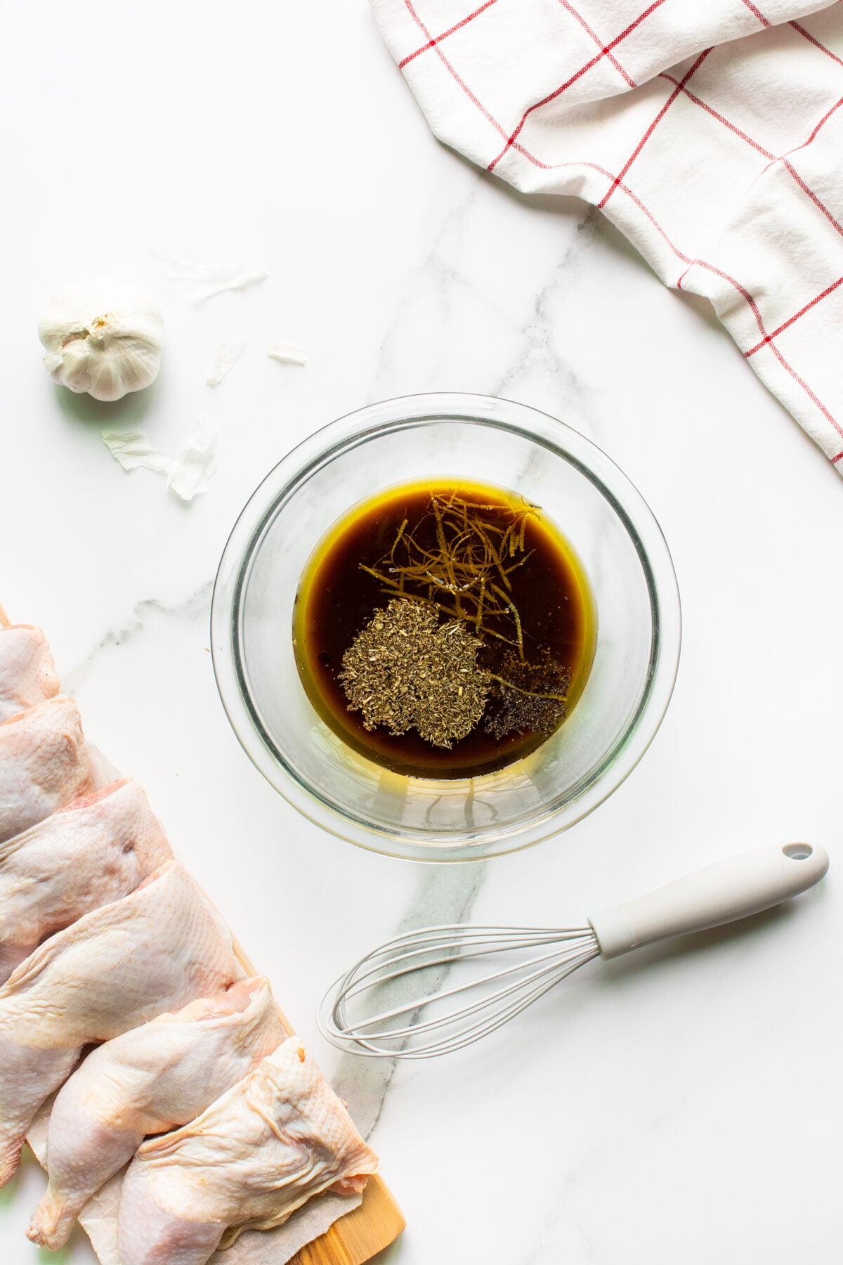 A glass bowl with soy sauce and herbs sits on a marble surface next to raw chicken wings, a whisk, a garlic bulb, and a white kitchen towel with red stripes.