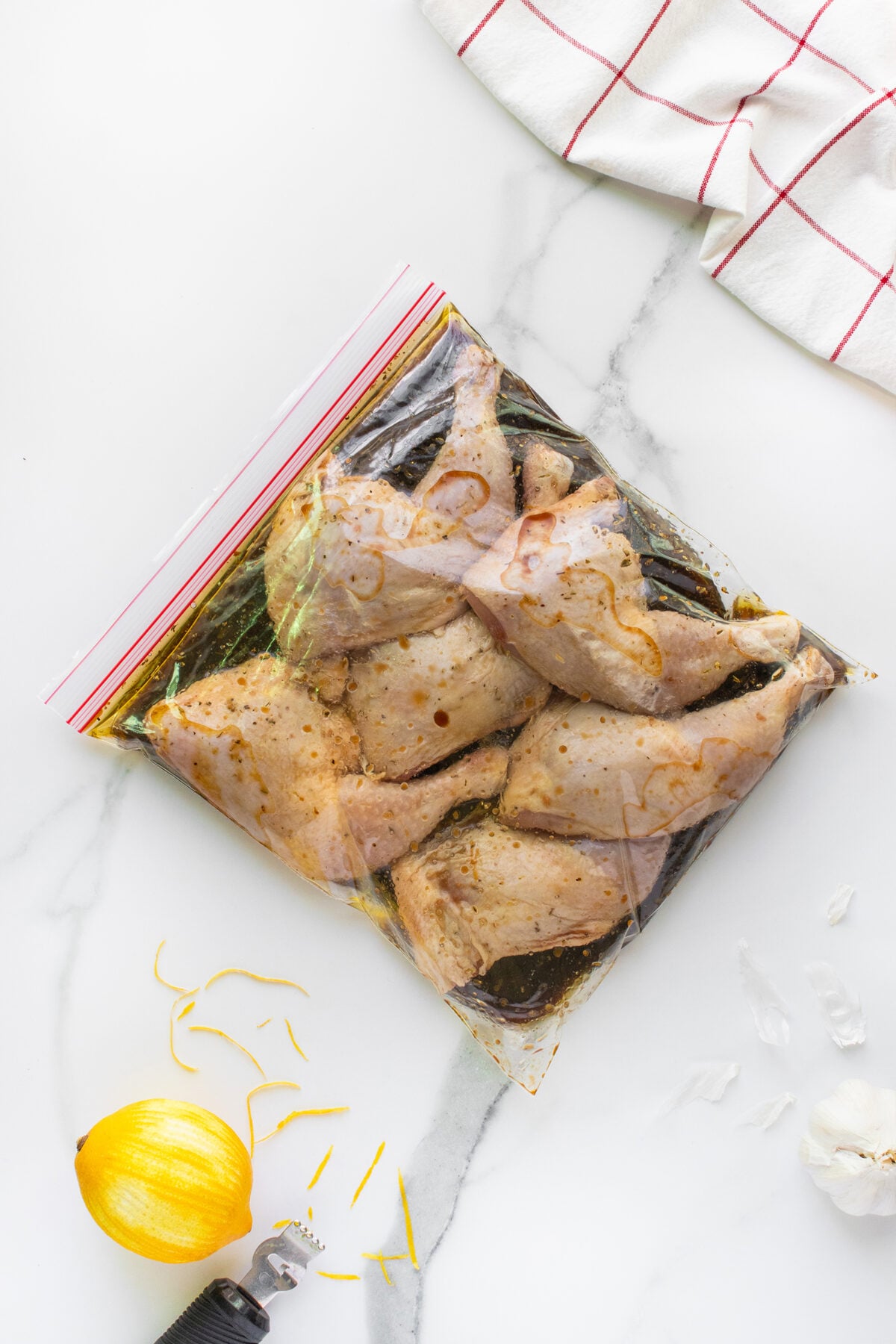 Raw chicken pieces marinating in a sealed plastic bag with herbs and liquid on a white marble surface, next to a zested lemon, lemon zest, garlic pieces, and a white towel with red stripes.