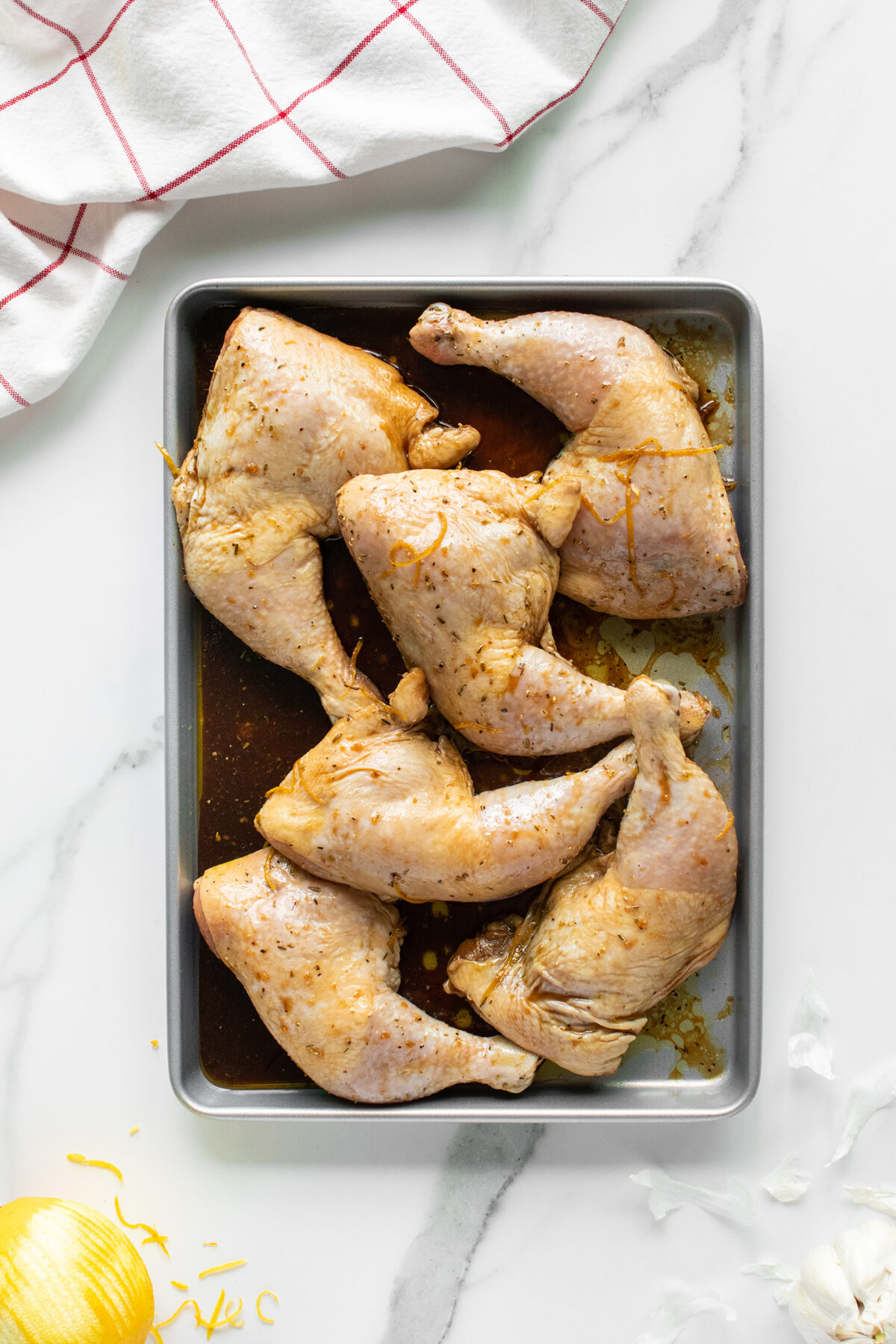 A metal baking tray with six marinated raw chicken leg quarters on a white marble surface, next to a red-and-white kitchen towel, lemon, and garlic cloves.