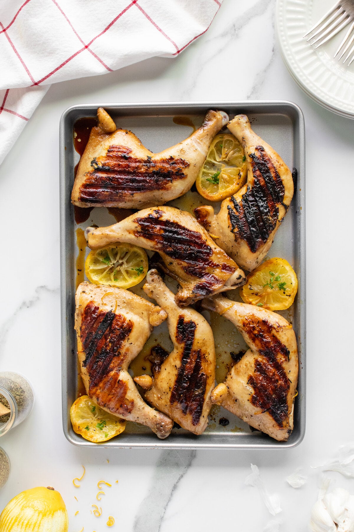 A baking tray with five grilled chicken leg quarters garnished with lemon slices, sitting on a white marble countertop next to a red-striped towel and stacked white plates.
