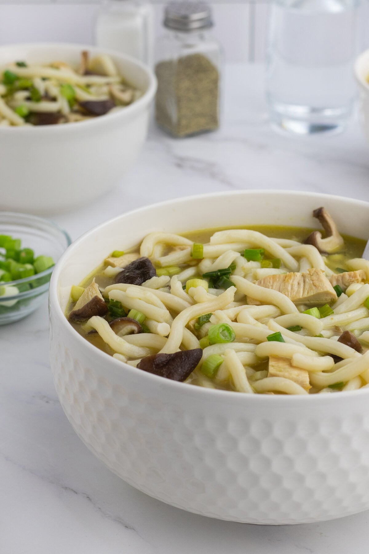 A white bowl filled with noodle soup, garnished with green onions and mushrooms, sits on a white surface. Another bowl, seasonings, and a small dish of chopped green onions are in the background.