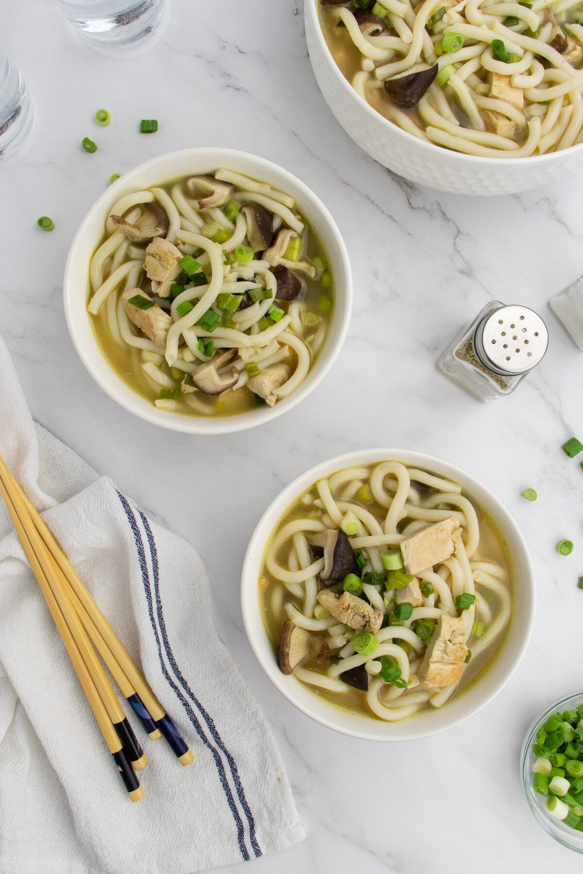 Three bowls of noodle soup with tofu, mushrooms, and green onions are on a white marble surface, alongside chopsticks, a salt shaker, a towel, and glasses of water. Scattered green onions garnish the table.