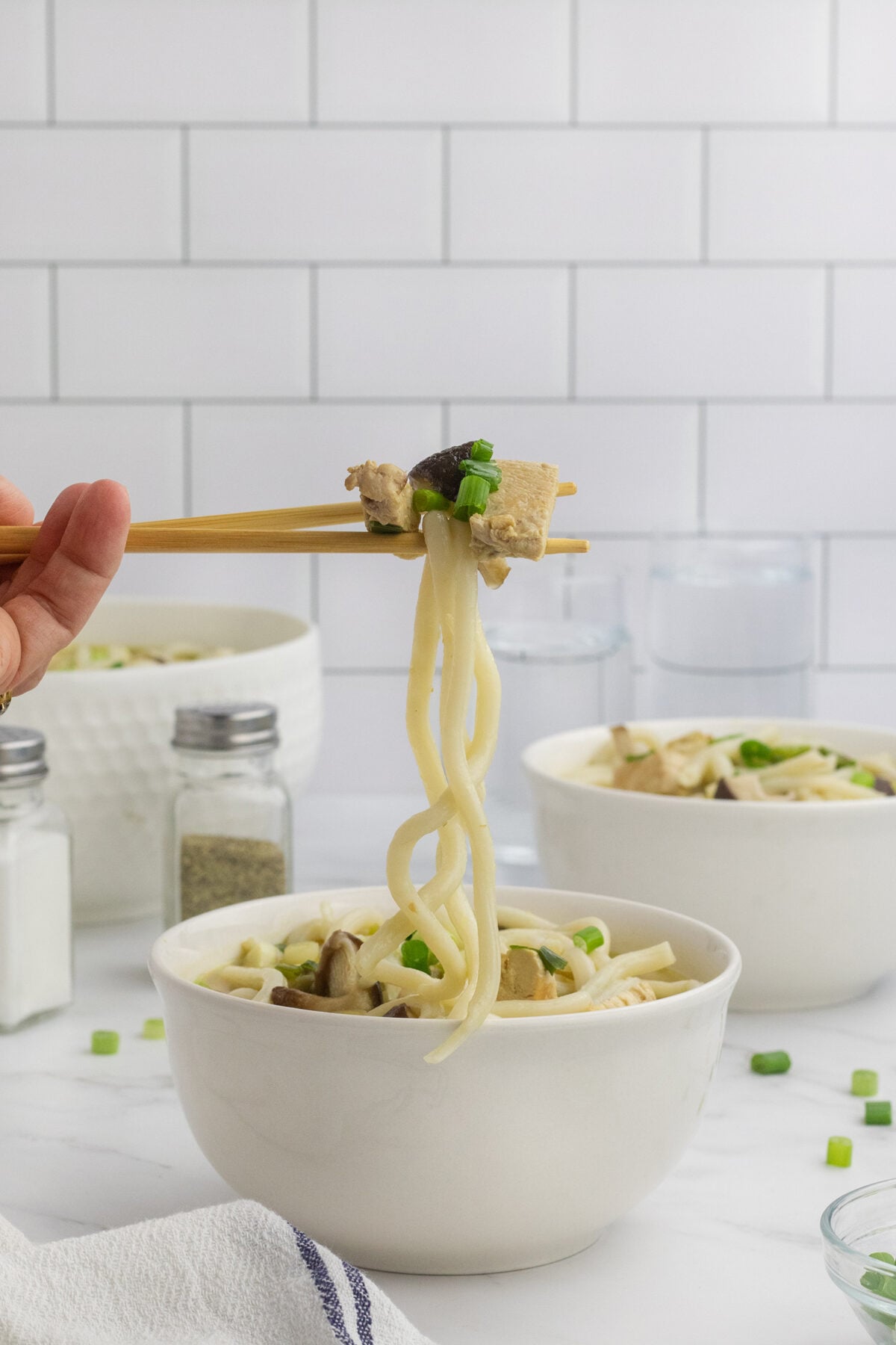 A hand holding chopsticks lifts noodles, tofu, and vegetables from a white bowl of soup, garnished with green onions. White tiles and kitchen items are visible in the background.