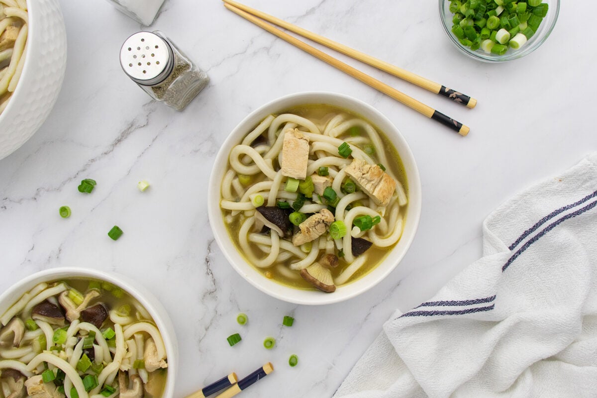 A bowl of noodle soup with tofu, mushrooms, and green onions sits on a white marble surface, surrounded by chopsticks, a pepper shaker, and a dish towel. Scattered green onions add color.
