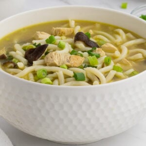 A white bowl filled with noodle soup containing pieces of chicken, mushrooms, and chopped green onions, set on a white surface with a small dish of extra green onions in the background.