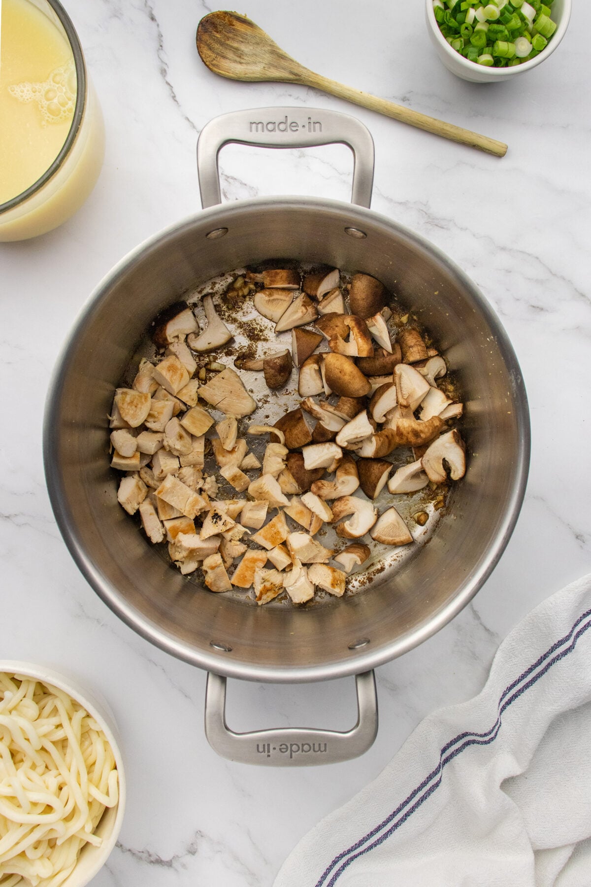 A stainless steel pot on a marble countertop contains chopped mushrooms being sautéed. Surrounding the pot are ingredients including broth, chopped green onions, a bowl of udon noodles, and a wooden spoon.