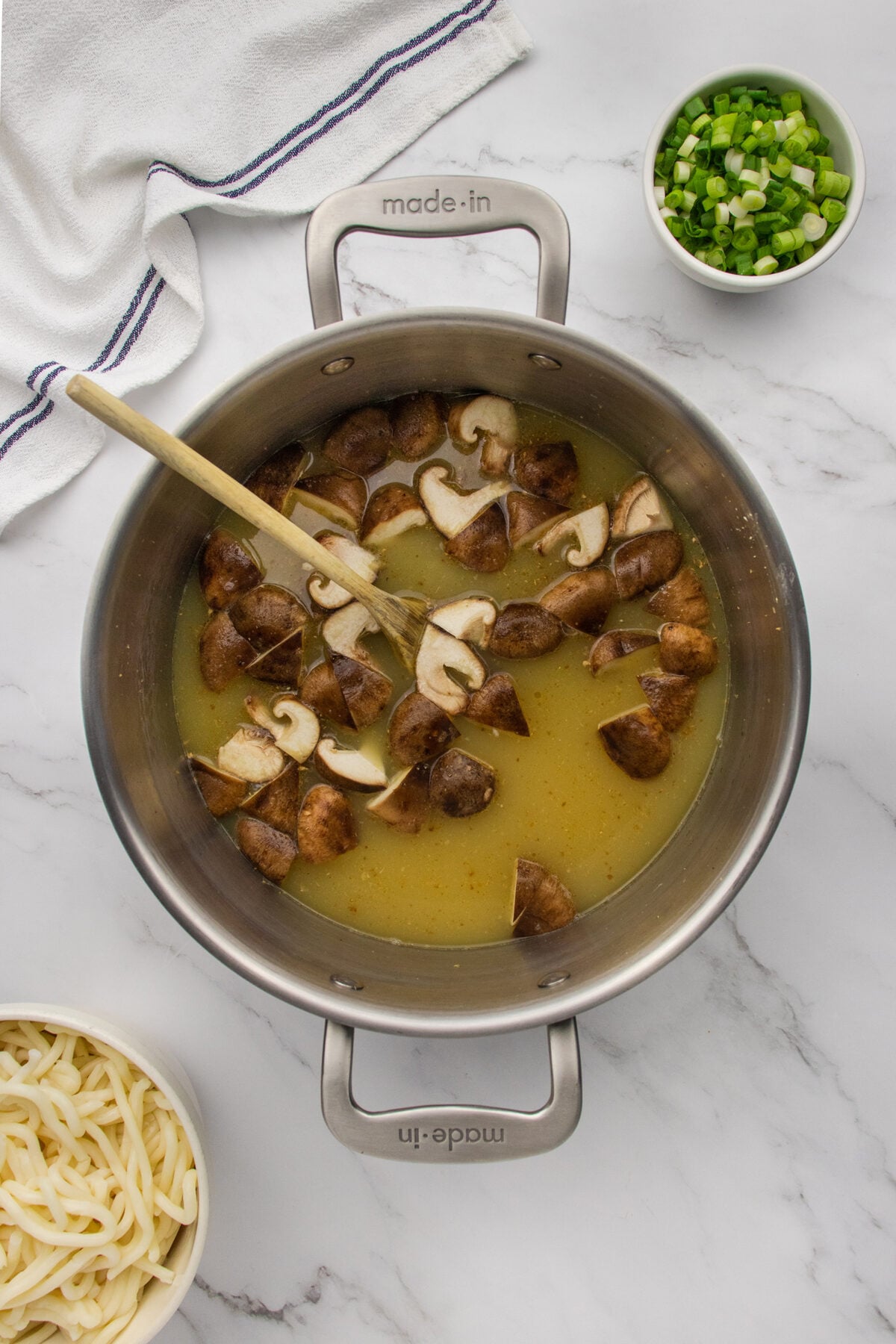 A metal pot with broth and sliced mushrooms being stirred with a wooden spoon, surrounded by bowls of chopped green onions and uncooked udon noodles on a white marble surface.