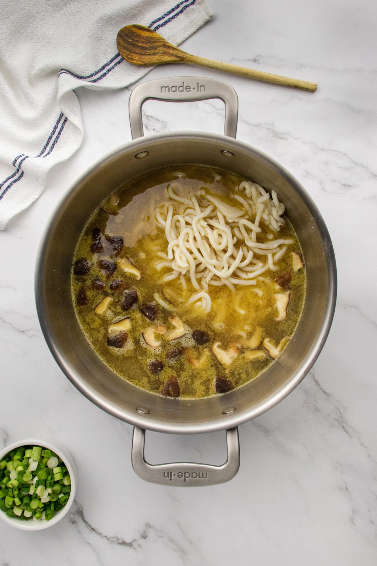 A metal pot filled with broth, sliced mushrooms, and udon noodles sits on a marble counter. Nearby are a bowl of chopped green onions, a wooden spoon, and a white towel with blue stripes.
