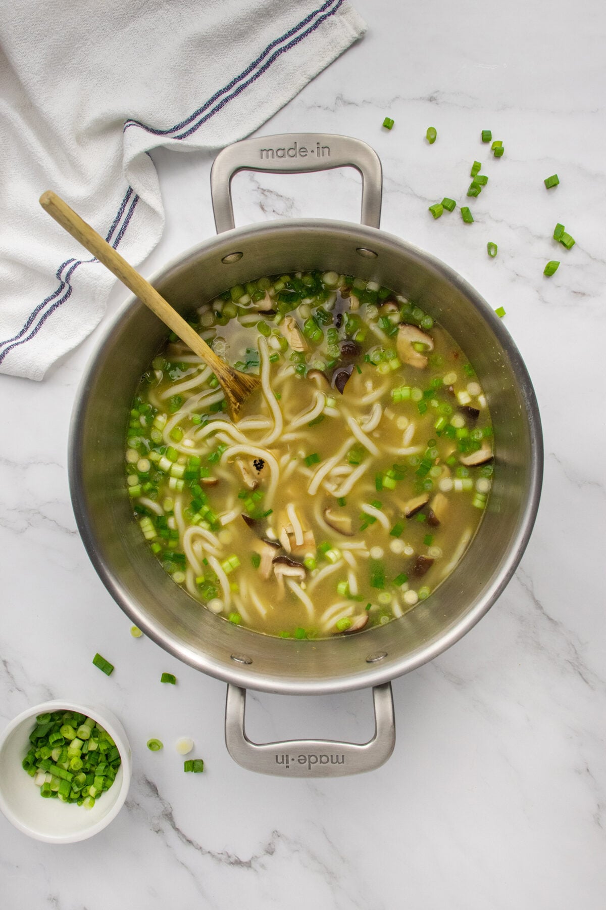 A pot of noodle soup with mushrooms and chopped green onions sits on a marble surface. A wooden spoon is in the pot, and green onions are scattered nearby. A bowl of green onions and a striped towel are also visible.