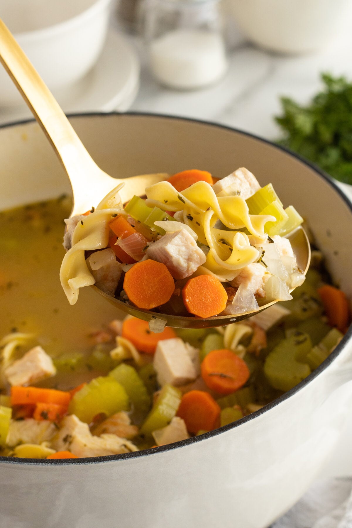 A ladle filled with homemade chicken noodle soup, featuring chunks of chicken, carrots, celery, and egg noodles, held over a large white pot containing more of the soup.