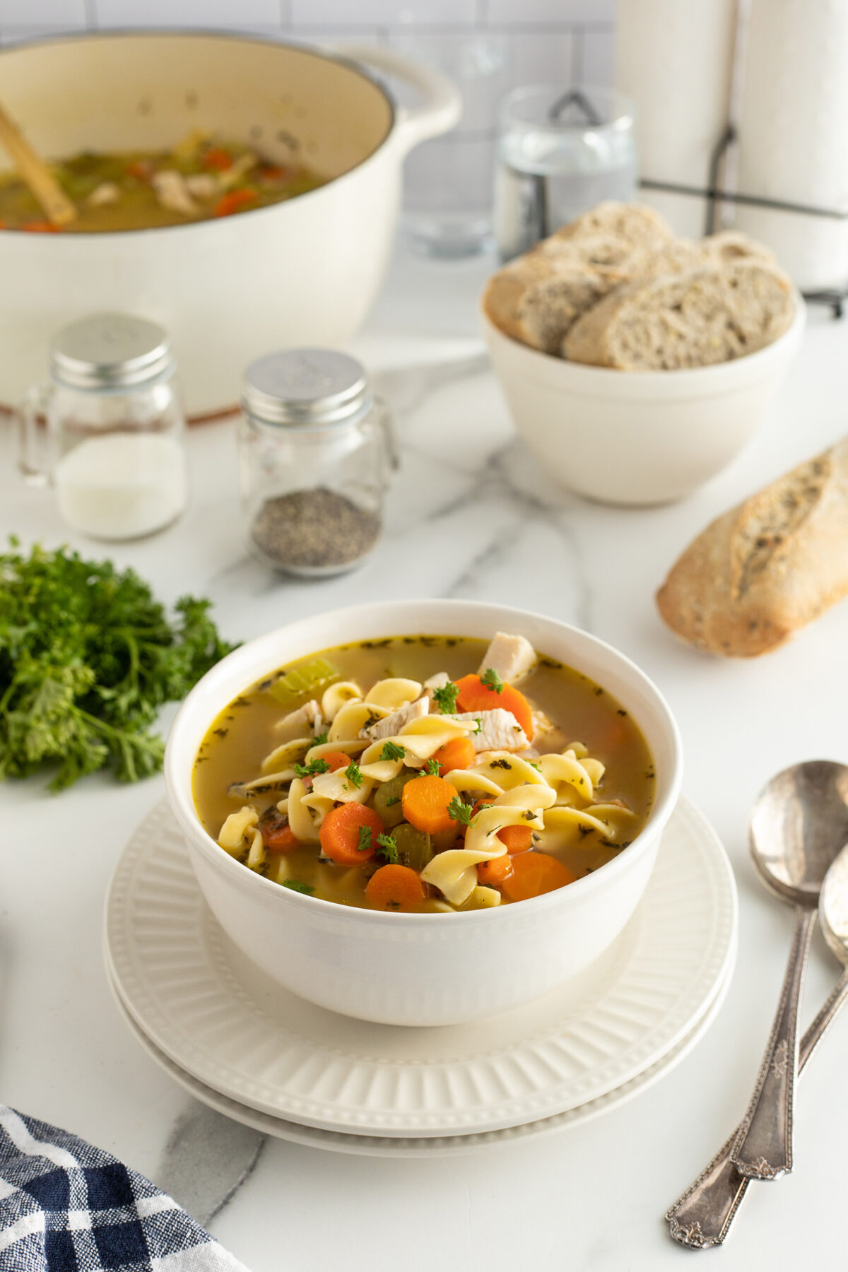 A white bowl of chicken noodle soup with carrots and noodles sits on a plate. In the background are a pot of soup, a bowl of bread, a baguette, salt and pepper shakers, fresh parsley, and two spoons.
