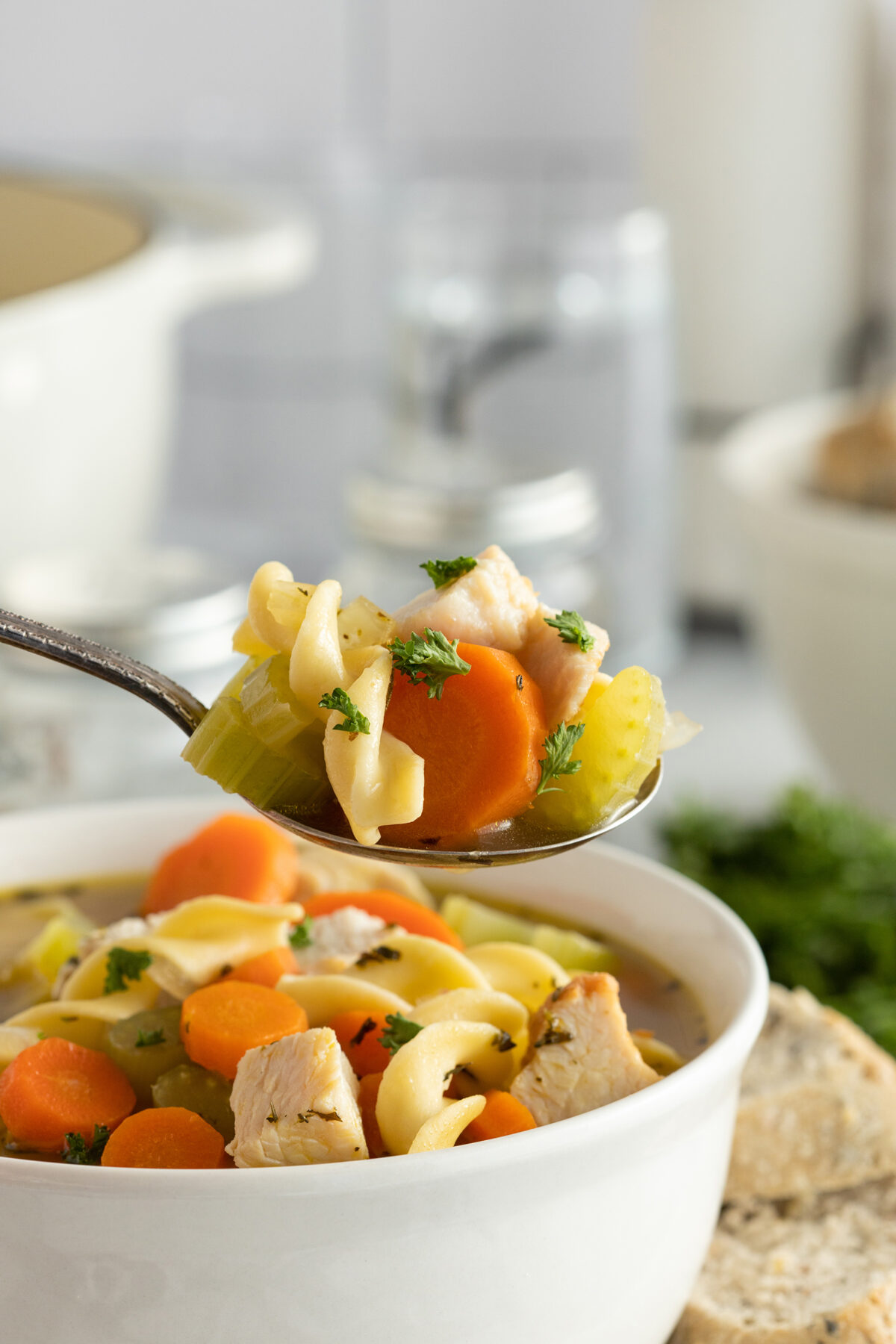 A spoonful of chicken noodle soup with carrots, celery, chicken, and noodles is held above a bowl filled with the same soup. Sliced bread is visible beside the bowl.