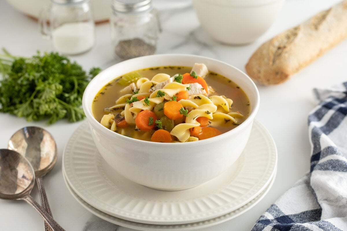 A white bowl filled with chicken noodle soup, featuring egg noodles, carrot slices, and herbs, sits on stacked plates. Fresh parsley, a baguette, and utensils are nearby on a white countertop.