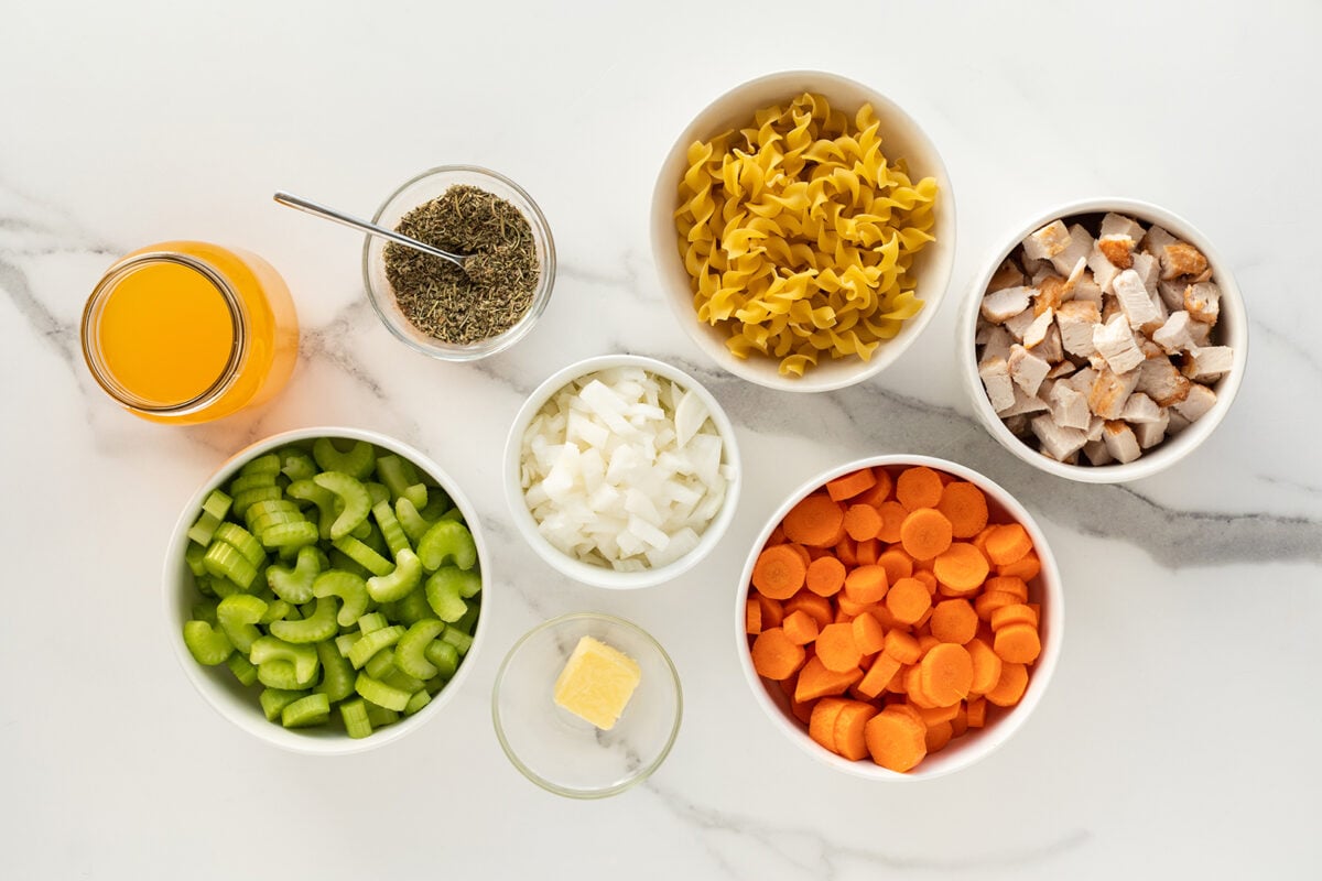 Overhead view of bowls containing chopped celery, onions, carrots, cooked chicken, dry pasta, dried herbs, a jar of broth, and a small dish with a pat of butter, arranged on a white marble surface.