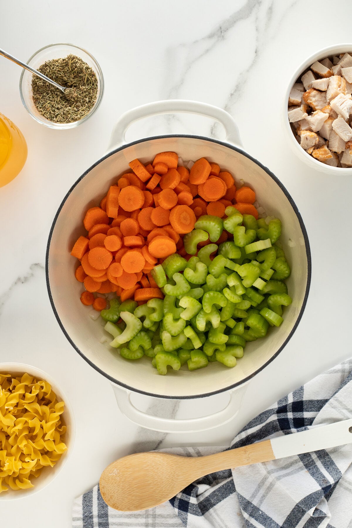 A white pot on a marble surface filled with sliced carrots and celery. Surrounding the pot are bowls of cubed chicken, dried herbs, egg noodles, and a jar of broth, with a wooden spoon and a striped towel nearby.