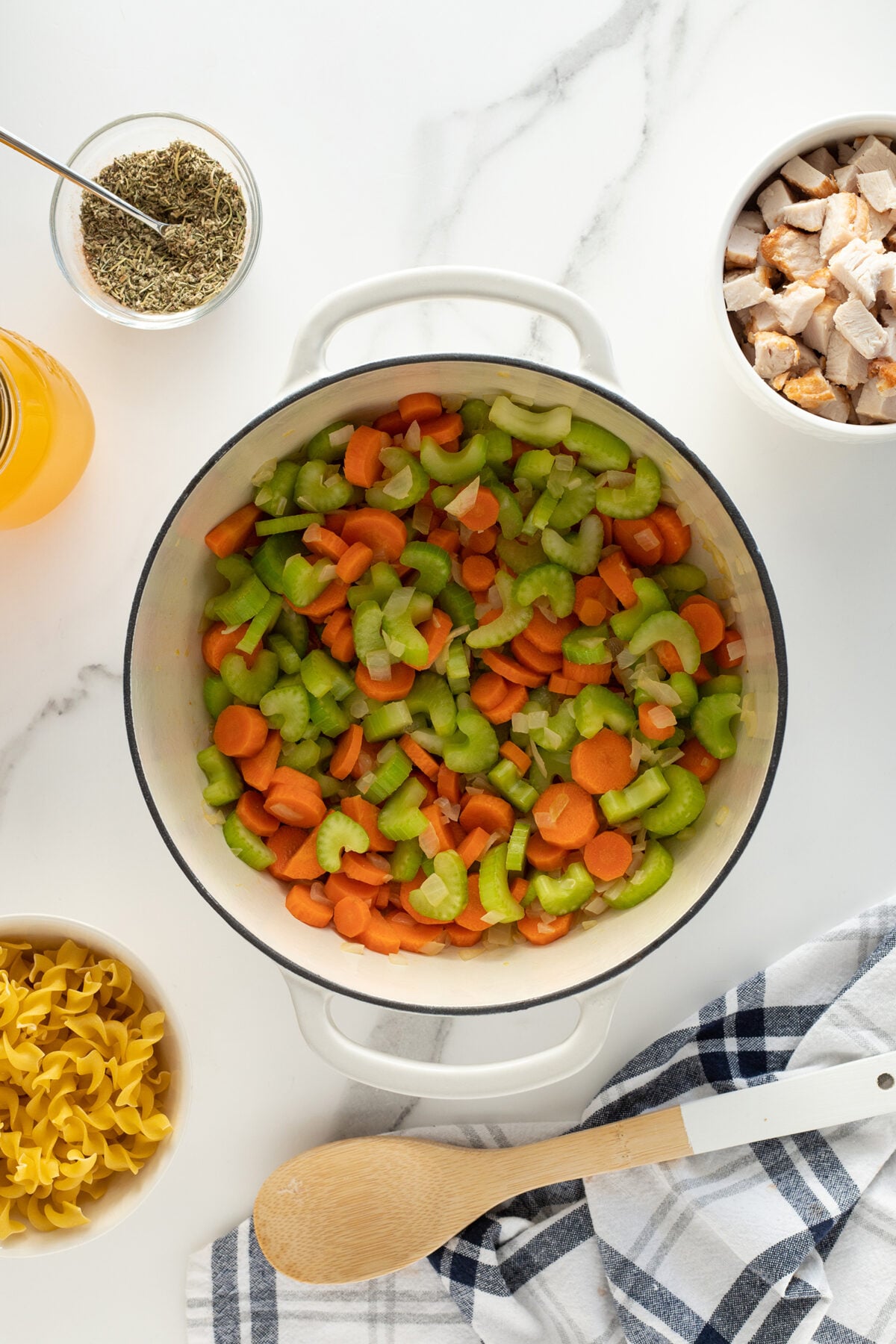 A pot filled with chopped celery, carrots, and onions sits on a white countertop surrounded by a bowl of dry pasta, a bowl of cooked chicken, a jar of broth, a bowl of herbs, and a wooden spoon on a striped towel.